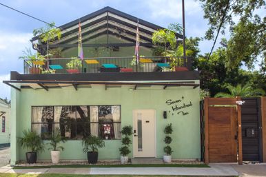 Mint-green two-story building with a rooftop patio featuring colorful umbrella shades, potted palms and string lights, white front door and wooden gate on a sunny neighborhood street.