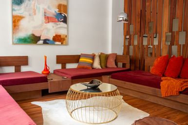 Cozy mid-century modern living room with low wooden seating, plush burgundy cushions, vibrant orange throw pillows, a gold wire round coffee table and abstract wall art.