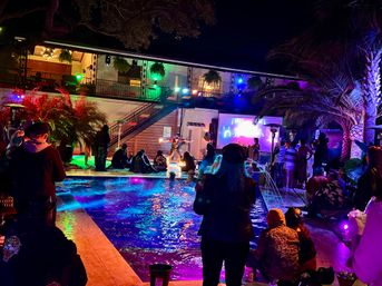 Nighttime outdoor pool party under palm trees with colorful neon lights, balcony venue, crowd gathered around a pool watching a fire performer and projection screen