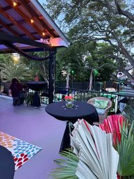 Twilight outdoor patio overlooking a poolside yard, string lights under a wooden pergola, cocktail tables with black linens and floral centerpieces, wicker seating with tropical cushions, palms and decorative fans for a festive tropical vibe.