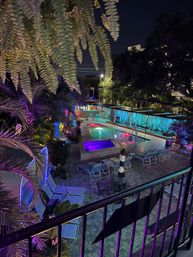 Neon-lit tropical outdoor pool and hot tub at night with palm trees, cabanas and lounge chairs on a tiled deck, glowing purple and teal lights