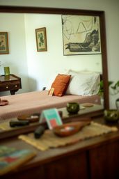 Cozy bedroom reflected in a mirror — pink quilted bed with white pillows and a rust-orange accent cushion, wooden dresser with small bowls and green glass decor, framed botanical and abstract art on a pale wall.