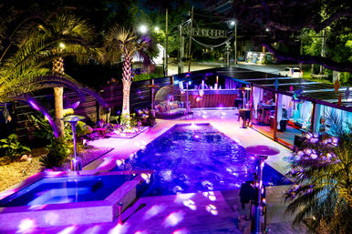 Resort-style backyard pool at night glowing purple, with adjacent hot tub, palm trees, cabana seating, outdoor bar and lit street beyond