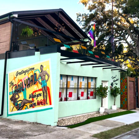 Mint-green two-story storefront with rooftop patio, playful retro postcard mural of swimsuit models, rainbow pride flag and potted trees on a tree-lined street.