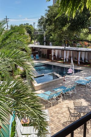 Resort-style outdoor pool and raised spa in a tropical courtyard with palm trees, paved sun deck, rows of blue lounge chairs, curtained cabanas and a pink pool lift under a sunny sky.