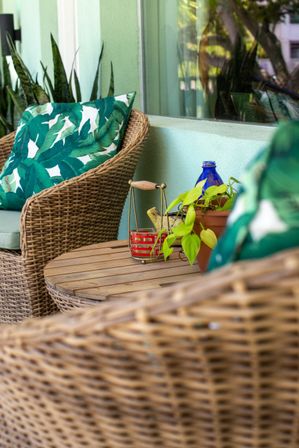 Cozy sunlit patio with woven wicker chairs and tropical green leaf cushions, round wooden side table holding a potted trailing plant, blue glass bottle and small red candle holder