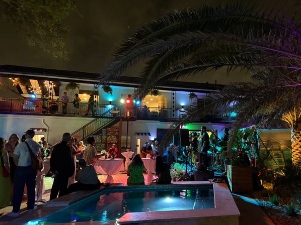 Poolside nighttime patio party in a tropical courtyard with palm trees, colorful lights and a live band on a small stage, guests mingling around cocktail tables under a two-story balcony.