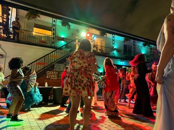 Nighttime courtyard dance party outside a two-story motel-style building with hanging plants, colorful stage lights, and a lively crowd in bright outfits.