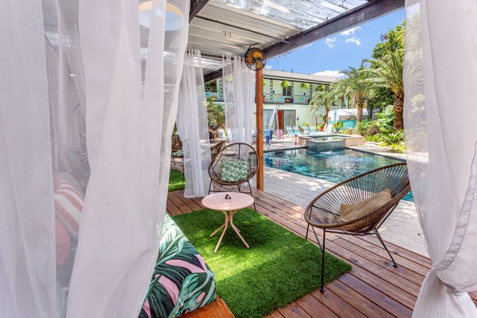 Sunlit poolside cabana with sheer white curtains, wicker lounge chairs and pink side table on green turf, overlooking a sparkling pool framed by palm trees and deck loungers