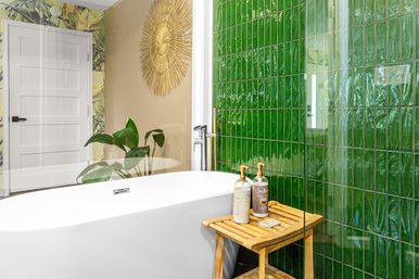 Modern bathroom with white freestanding bathtub, glossy green tiled wall, wooden stool with bath products, tropical plant and sunburst wall decor