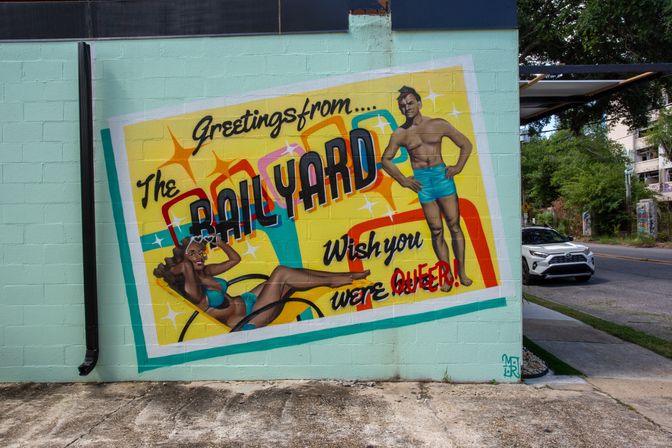 Colorful postcard-style mural on a mint-green cinderblock wall at a street corner, featuring a reclining woman and standing man in vintage swimwear, retro stars and bold lettering that reads 'Greetings from...' and 'Wish you were QUEER!', with a sidewalk and parked white SUV visible nearby.