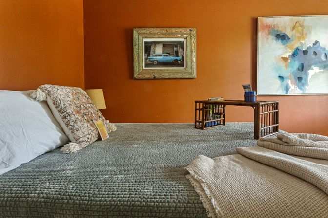 Cozy bedroom with a warm orange accent wall, textured gray bedspread and woven throw, patterned decorative pillow, framed vintage car photo, abstract wall art, and a low wooden bed tray holding books and a cup