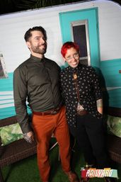 Two smiling adults posing in front of a turquoise retro camper backdrop at an outdoor event — man in olive shirt and rust pants, person with short red hair, dotted face makeup, floral black shirt and bolo tie.