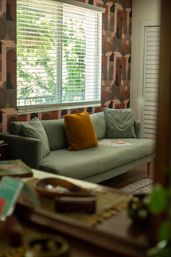 Cozy modern living room with a green-gray sofa, mustard accent pillow, geometric wallpaper and a sunlit window with blinds revealing green trees outside.