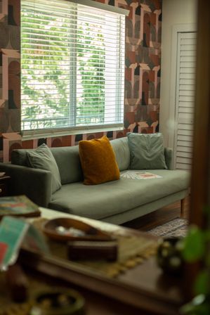 Cozy modern living room with a green-gray sofa, mustard accent pillow, geometric wallpaper and a sunlit window with blinds revealing green trees outside.