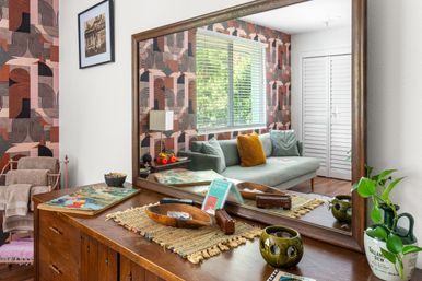 Sunlit living room interior with retro geometric pink and brown wallpaper, wooden dresser topped with bowls, plants and a woven runner, and a large mirror reflecting a green sofa with a mustard cushion and window blinds