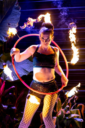 Daring fire performer spinning a flaming hula hoop during a nighttime outdoor show surrounded by tropical foliage