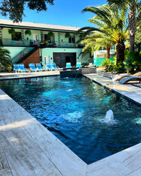 Sunlit palm-lined poolside oasis with bubbling fountain, turquoise lounge chairs, hanging ferns and a two-story retro-style courtyard building — tropical staycation vibe