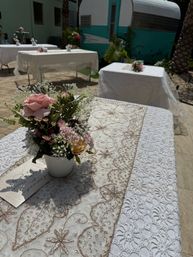 Sunlit outdoor patio reception tables with white lace tablecloths and beaded gold-embroidered runners, small pink rose and baby’s-breath floral centerpiece in a white pot and a turquoise vintage camper in the background