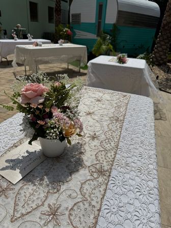 Sunlit outdoor patio reception tables with white lace tablecloths and beaded gold-embroidered runners, small pink rose and baby’s-breath floral centerpiece in a white pot and a turquoise vintage camper in the background