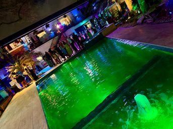 Nighttime outdoor pool party with neon-green illuminated water, guests gathered along the pool deck and upper balcony, colorful uplighting on palm trees and a modern two-story building