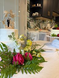 Two-tier white wedding cake topped with a bride-and-groom figurine under a bell, surrounded by pink roses, yellow blooms, ferns and lavender on a decorated kitchen table with a berry-topped cake and patterned backsplash in the background.