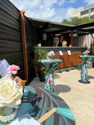 Sunny outdoor patio bar with wooden counter, large beverage dispensers and tropical-print cocktail tables tied with floral accents for a casual daytime event.