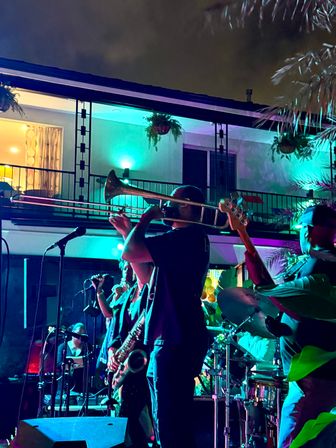 Live music on an outdoor patio at night — brassy trombone player fronting a band with saxophone, bass and drums under teal and purple stage lights and a balcony with hanging plants