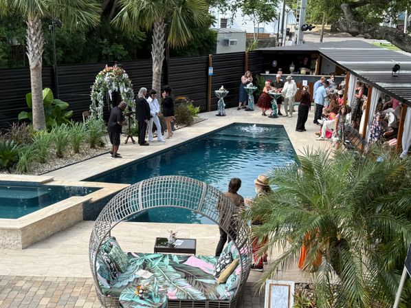 Tropical backyard poolside wedding reception with guests mingling by a floral arch, palm trees, cabana bar and a wicker daybed with bold leaf-print cushions.