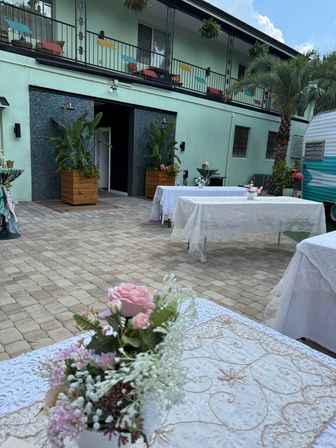 Outdoor tropical courtyard event setup with lace-covered tables and pink floral centerpieces on stone pavers, mint-green two-story building with balcony and colorful flags, large planters, palm tree and a retro teal camper in the background.