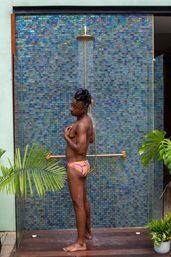 Person enjoying a resort-style outdoor shower under a brass rain showerhead, standing in bright patterned swim briefs against an iridescent blue mosaic tile wall with tropical plants nearby.