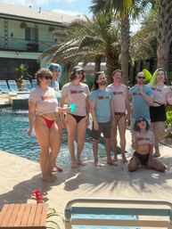 Group of eight adults in matching cropped tees and swimwear posing and laughing poolside in a sunny tropical courtyard with palm trees, lounge chairs and a two-story balcony in the background.