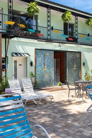 Sunny courtyard patio with mosaic-tiled outdoor showers, white sun loungers and blue-striped chairs, hanging ferns and a second-floor balcony with colorful geometric accents.