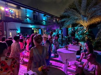 Vibrant outdoor nighttime party on a tropical patio with a live band on stage, colorful purple and blue uplighting, palm trees, cocktail tables with flowers, and guests dancing and mingling.