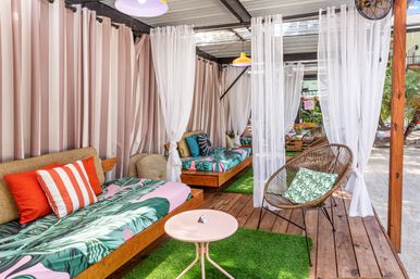 Cozy tropical outdoor cabana lounge on a wooden deck with patterned daybeds, bold throw pillows, sheer white curtains and a rattan chair.