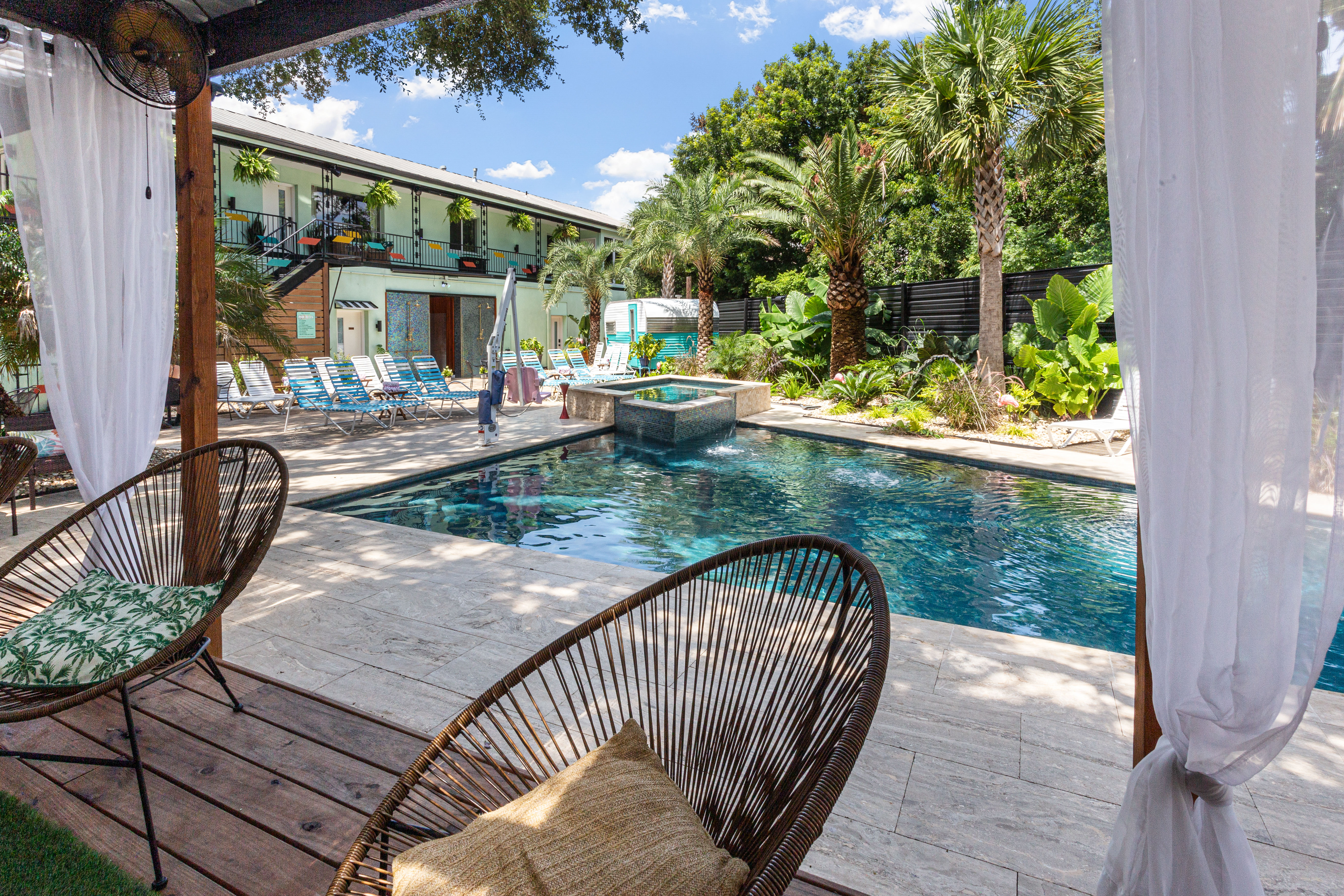 Resort-style outdoor pool courtyard with turquoise water, palm trees, lined lounge chairs and a cabana framed by sheer curtains and wicker seating.