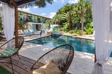 Resort-style outdoor pool courtyard with turquoise water, palm trees, lined lounge chairs and a cabana framed by sheer curtains and wicker seating.