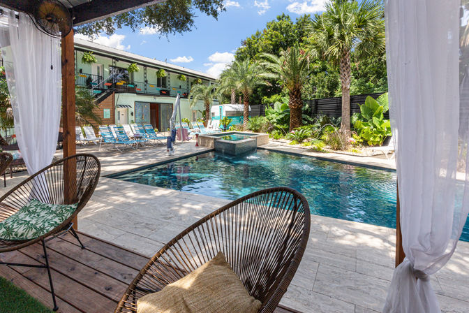 Resort-style outdoor pool courtyard with turquoise water, palm trees, lined lounge chairs and a cabana framed by sheer curtains and wicker seating.