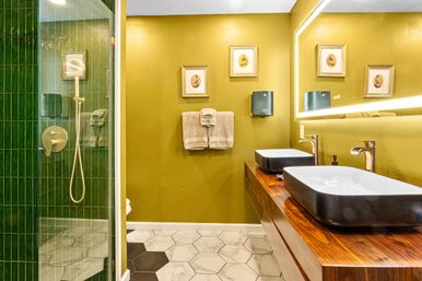 Modern bathroom with olive-yellow walls, backlit mirror, double white vessel sinks on a walnut vanity, green-tiled glass shower and hexagon floor tiles.