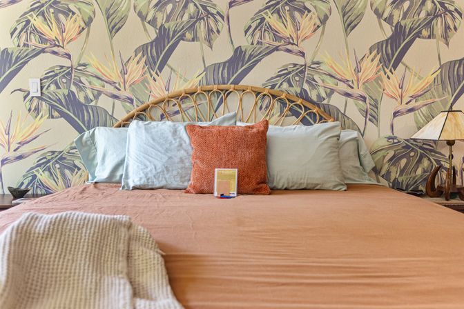 Cozy tropical bedroom interior with woven rattan headboard, botanical bird-of-paradise wallpaper, rust accent pillow, sage pillows, terracotta bedding and knit throw.