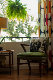 Cozy mid-century modern reading nook with a green upholstered armchair topped by a whimsical skeletal tuxedo pillow, a hanging fern by a sunlit balcony window, wooden side table with potted plant and retro lamp, and bold geometric curtains.