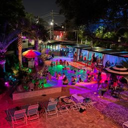 Vibrant nighttime outdoor pool party with multicolored LED lights, palm trees, cabanas and lounge chairs, groups socializing in and around an illuminated urban pool