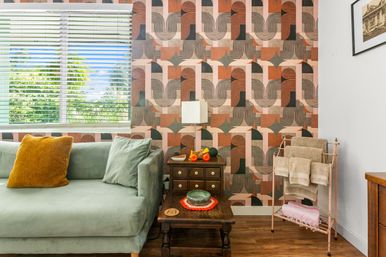 Bright living room with seafoam-green velvet sofa and mustard pillow, bold retro geometric pink-and-terracotta wallpaper, wooden side table with fruit and bowl, towel rack, and a window with blinds showing lush tropical greenery outside.