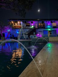 Vibrant nighttime outdoor pool with arcing water jets and colorful purple, blue and green LED lights reflecting on the water, two-story balcony with lounge chairs and hanging plants under a crescent moon.