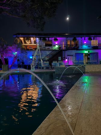 Vibrant nighttime outdoor pool with arcing water jets and colorful purple, blue and green LED lights reflecting on the water, two-story balcony with lounge chairs and hanging plants under a crescent moon.