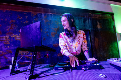 Smiling DJ spinning tracks on a laptop and controller in an urban nightclub with purple-green lighting and a textured metal wall backdrop.