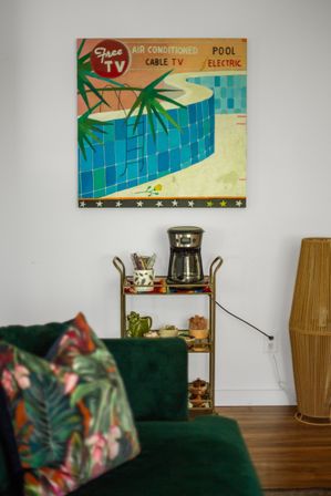 Cozy living room corner with retro pool wall art above a gold bar-cart coffee station, dark green velvet sofa with tropical-print pillows, wicker floor lamp and hardwood floors