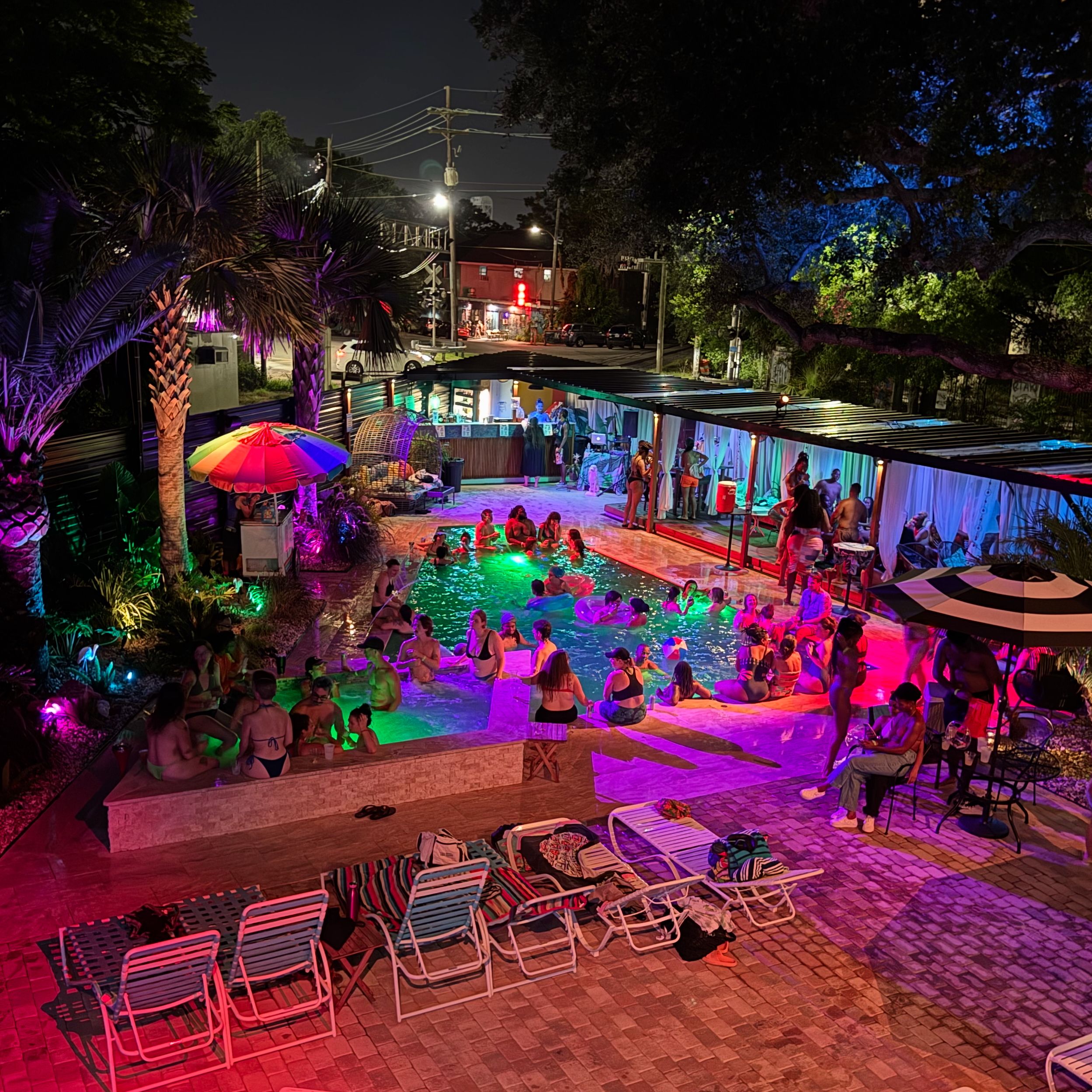 Vibrant nighttime outdoor pool party with multicolored LED lighting, people swimming and lounging, palm trees, cabanas and umbrellas, lounge chairs on a brick deck, and an urban street visible beyond.