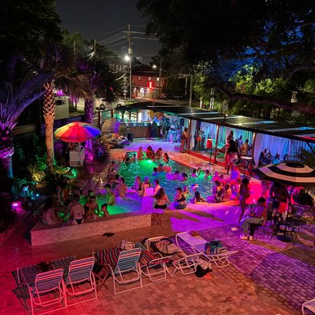Vibrant nighttime outdoor pool party with multicolored LED lighting, people swimming and lounging, palm trees, cabanas and umbrellas, lounge chairs on a brick deck, and an urban street visible beyond.