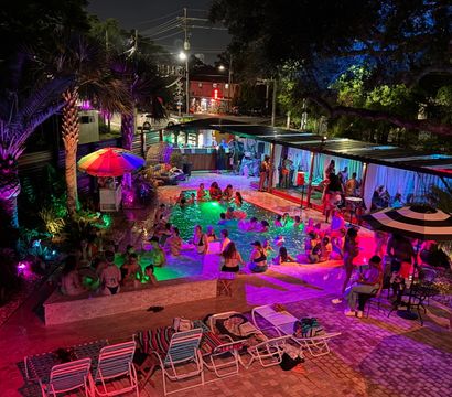 Vibrant nighttime outdoor pool party with multicolored LED lighting, people swimming and lounging, palm trees, cabanas and umbrellas, lounge chairs on a brick deck, and an urban street visible beyond.